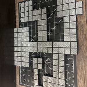 Op de afbeelding: Grijze en zwarte in elkaar grijpende bouwstenen gerangschikt op een zwarte snijmat met inch-metingen. De blokken hebben een oppervlak met textuur dat op bakstenen lijkt. De opstelling suggereert een bouw- of ontwerpproject.