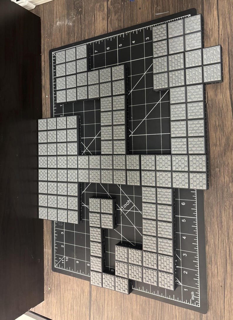 Op de afbeelding: Grijze en zwarte in elkaar grijpende bouwstenen gerangschikt op een zwarte snijmat met inch-metingen. De blokken hebben een oppervlak met textuur dat op bakstenen lijkt. De opstelling suggereert een bouw- of ontwerpproject.