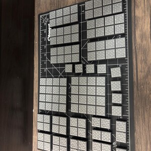 Op de afbeelding: Een verzameling grijze, getextureerde, modulaire terreinstukken gerangschikt op een zwarte snijmat met meetmarkeringen. De stukken zijn in verschillende maten en vormen, ontworpen voor tabletop gaming.