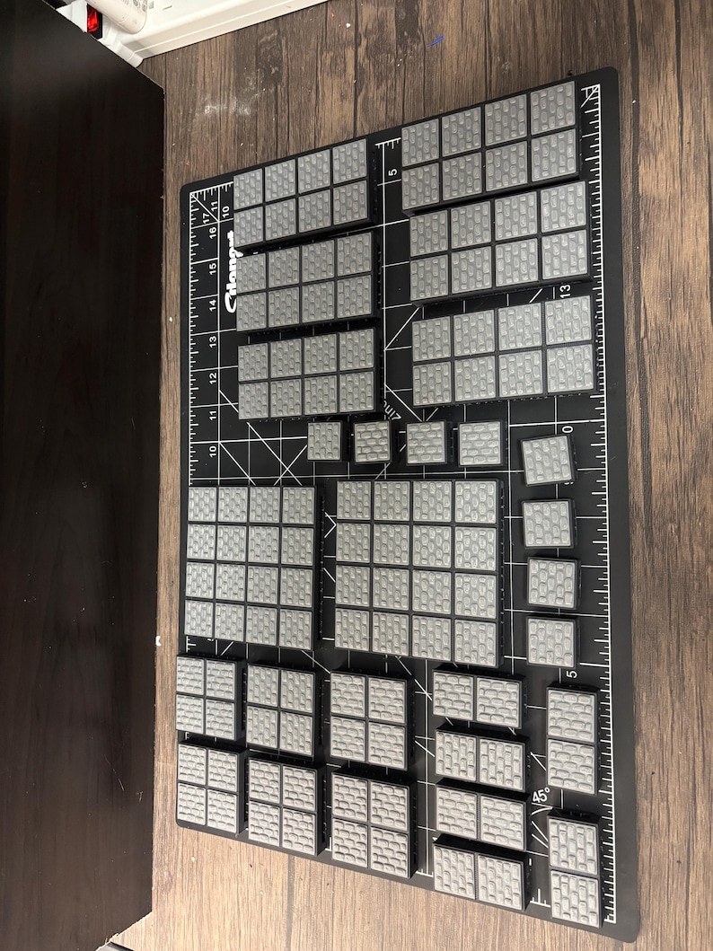 Op de afbeelding: Een verzameling grijze, getextureerde, modulaire terreinstukken gerangschikt op een zwarte snijmat met meetmarkeringen. De stukken zijn in verschillende maten en vormen, ontworpen voor tabletop gaming.