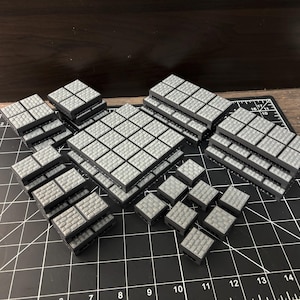 Op de afbeelding: Een verzameling grijze, vierkante, modulaire terreinstukken voor tabletop gaming. De stukken zijn in verschillende configuraties gestapeld en tonen een getextureerd, baksteenachtig oppervlak. De stukken zijn gerangschikt op een zwarte snijmat met witte rasterlijnen.