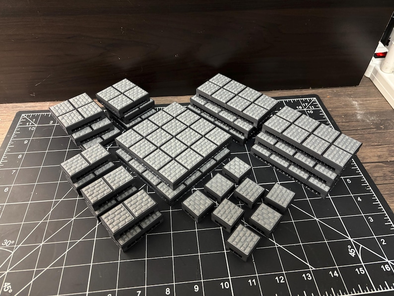 Op de afbeelding: Een verzameling grijze, vierkante, modulaire terreinstukken voor tabletop gaming. De stukken zijn in verschillende configuraties gestapeld en tonen een getextureerd, baksteenachtig oppervlak. De stukken zijn gerangschikt op een zwarte snijmat met witte rasterlijnen.