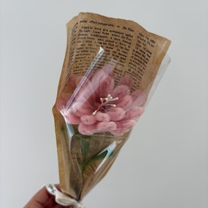 May include: A bouquet of pink flowers wrapped in brown paper with newspaper print and clear cellophane. The bouquet is tied with a white ribbon. The flowers have a soft, textured appearance.