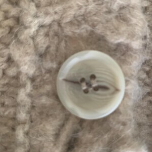 May include: Close-up of a round, off-white button with four holes, attached to a textured, beige fabric. The button has a subtle, pearlescent sheen, and the fabric appears soft and fuzzy.