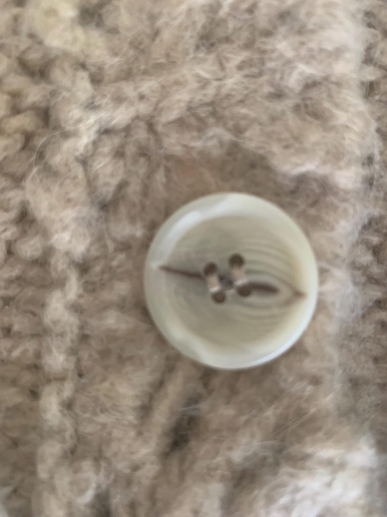 May include: Close-up of a round, off-white button with four holes, attached to a textured, beige fabric. The button has a subtle, pearlescent sheen, and the fabric appears soft and fuzzy.