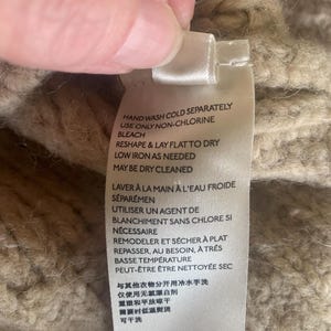 May include: Close-up of a clothing tag with care instructions in English and French. The tag is white with black text, and the background is a textured, beige knit fabric. Instructions include hand washing, non-chlorine bleach, and low iron.