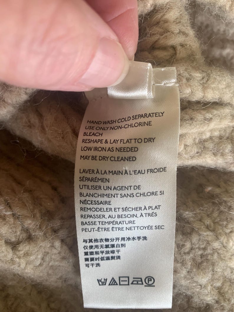 May include: Close-up of a clothing tag with care instructions in English and French. The tag is white with black text, and the background is a textured, beige knit fabric. Instructions include hand washing, non-chlorine bleach, and low iron.