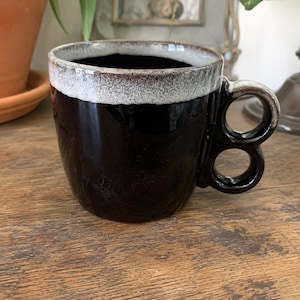 May include: A dark brown ceramic mug featuring a wide, textured white rim and a unique handle design composed of two stacked circular loops. The body of the mug has a glossy finish, while the rim and handle edges show a speckled, matte texture.