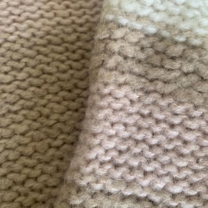 May include: Close-up of a knitted textile in shades of beige, cream, and light brown. The chunky knit pattern is visible, creating a textured surface. The fabric appears soft and warm, suggesting a cozy garment or home decor item.