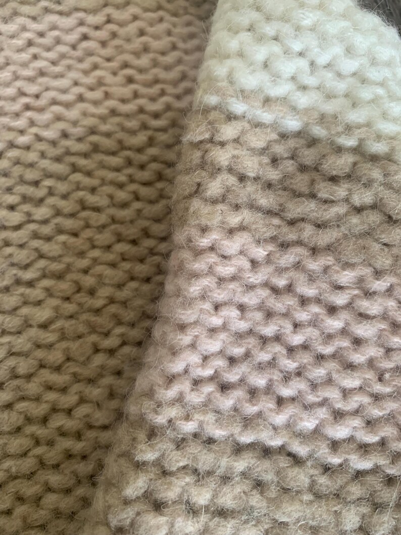 May include: Close-up of a knitted textile in shades of beige, cream, and light brown. The chunky knit pattern is visible, creating a textured surface. The fabric appears soft and warm, suggesting a cozy garment or home decor item.