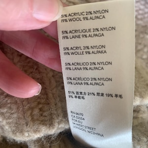 May include: Close-up of a beige knit garment with a tag. The tag lists the fabric composition as 51% acrylic, 21% nylon, 19% wool, and 9% alpaca. Additional text includes the garment's RN and CA numbers, and the address of the manufacturer.