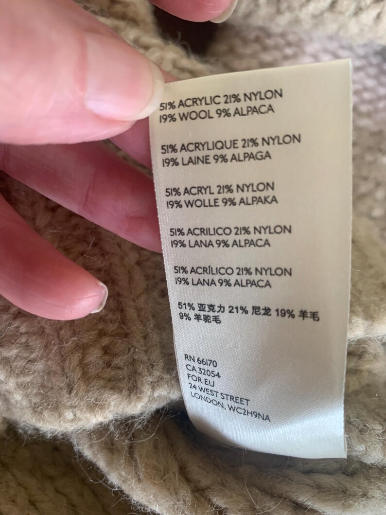 May include: Close-up of a beige knit garment with a tag. The tag lists the fabric composition as 51% acrylic, 21% nylon, 19% wool, and 9% alpaca. Additional text includes the garment's RN and CA numbers, and the address of the manufacturer.