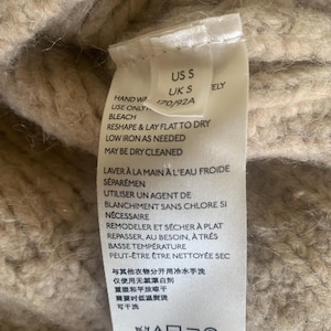 May include: Close-up of a beige knit garment tag with care instructions. The tag includes text in English and French, with washing and drying symbols. The garment size is US S and UK S.