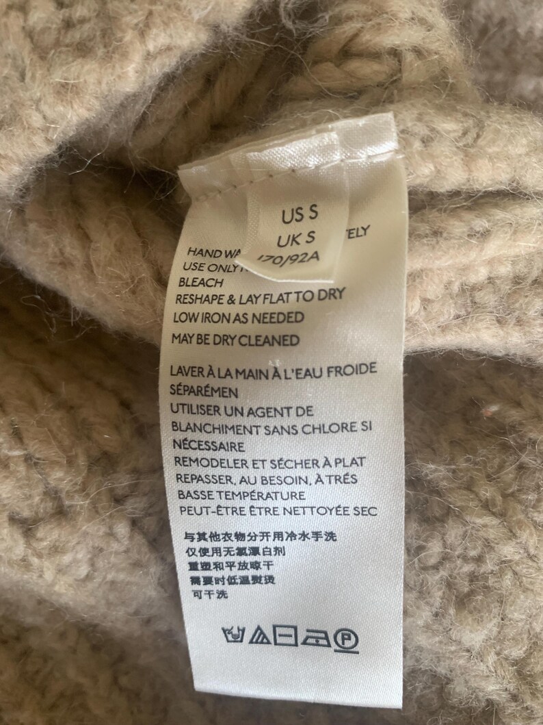 May include: Close-up of a beige knit garment tag with care instructions. The tag includes text in English and French, with washing and drying symbols. The garment size is US S and UK S.