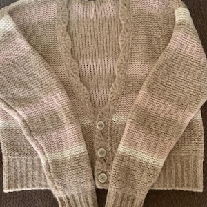 May include: A beige, pink, and white striped cardigan sweater with a v-neckline and three round buttons. The sweater has long sleeves and a textured knit design.