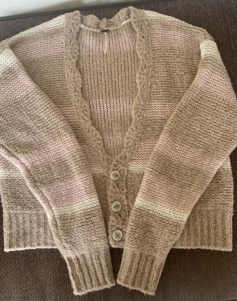 May include: A beige, pink, and white striped cardigan sweater with a v-neckline and three round buttons. The sweater has long sleeves and a textured knit design.