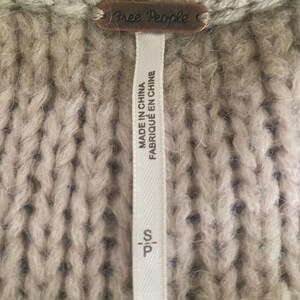 May include: Close-up of a beige knit sweater with a Free People brand tag. The tag is a small, rectangular metal plate. A white tag with the text "MADE IN CHINA FABRIQUE EN CHINE" is attached to the sweater.