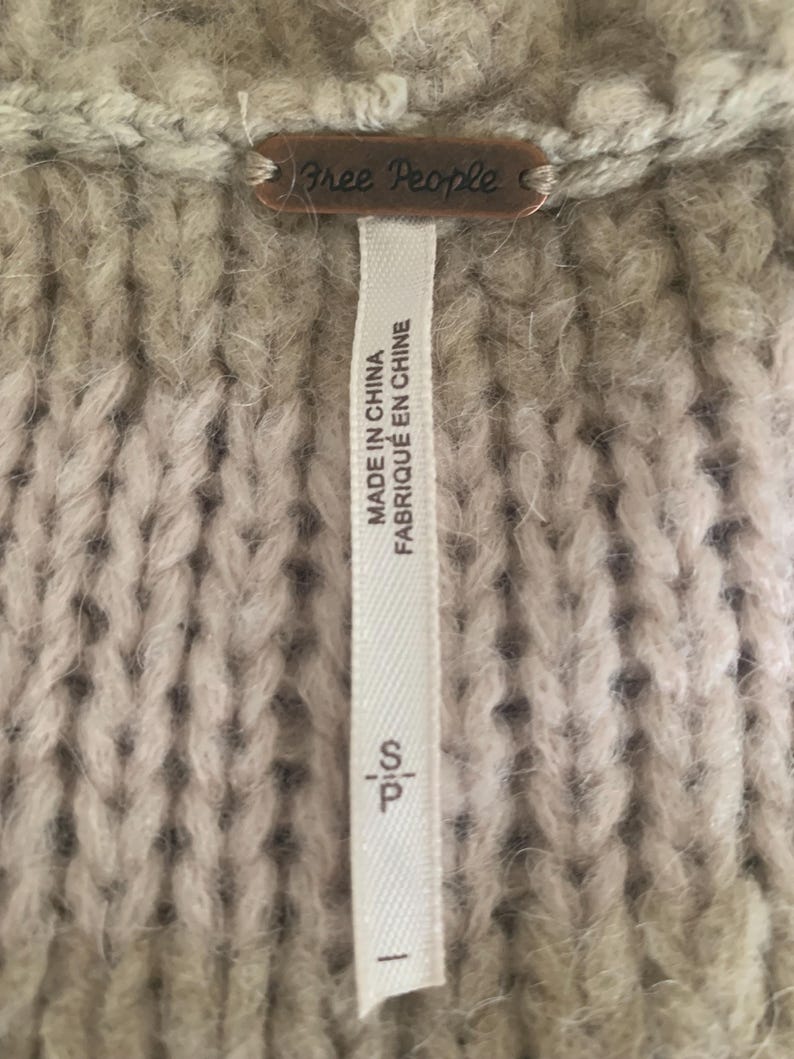 May include: Close-up of a beige knit sweater with a Free People brand tag. The tag is a small, rectangular metal plate. A white tag with the text "MADE IN CHINA FABRIQUE EN CHINE" is attached to the sweater.