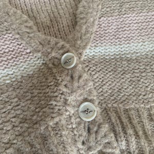 May include: Close-up of a beige cardigan with three white buttons. The knitted sweater has a V-neck and a striped pattern in shades of beige, pink, and white. The buttons are round with a decorative design.