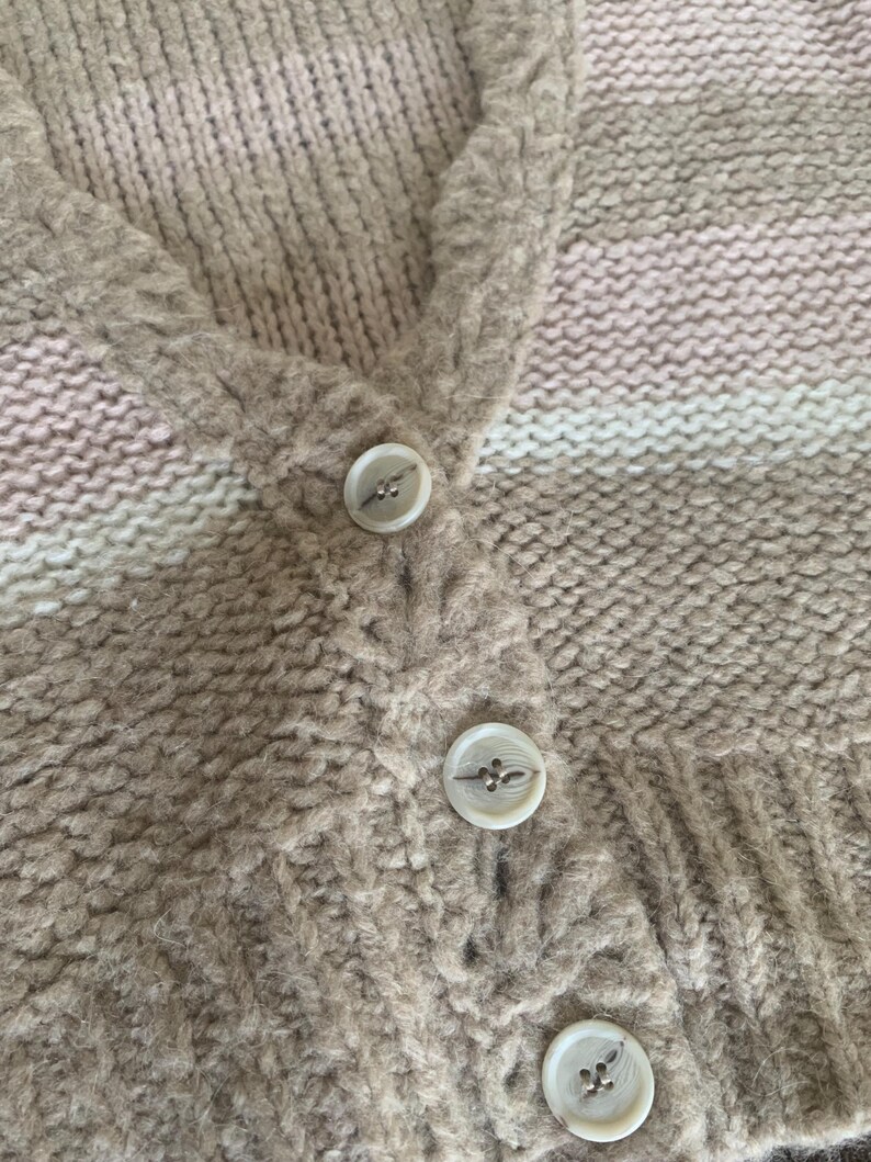 May include: Close-up of a beige cardigan with three white buttons. The knitted sweater has a V-neck and a striped pattern in shades of beige, pink, and white. The buttons are round with a decorative design.