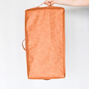 Puede incluir: Una funda de ropa rectangular de color naranja con cierre de cremallera y asa. La bolsa es sostenida por una mano sobre un fondo blanco. La bolsa tiene una superficie texturizada.