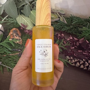 May include: A clear glass bottle of face serum with a wooden cap. The label reads "Vitamin C + Hyaluronic Face Serum" and lists ingredients. The bottle contains 2 oz / 60 ml of serum. The Alchemy of Life is printed at the bottom of the label.