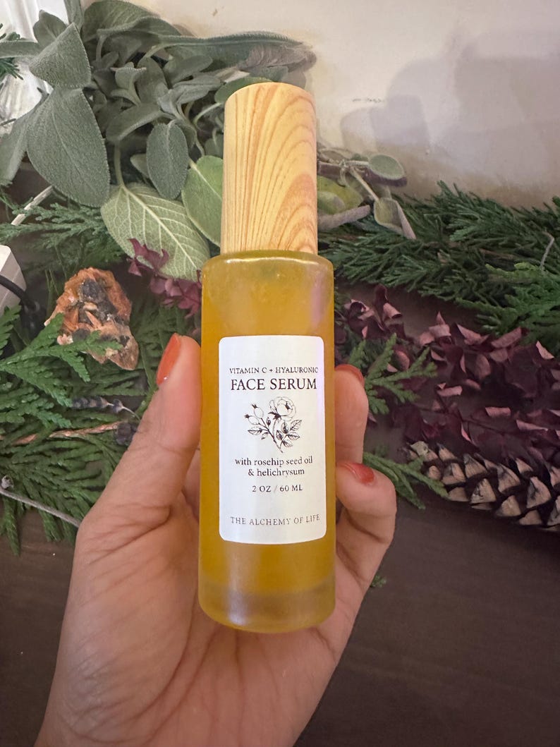 May include: A clear glass bottle of face serum with a wooden cap. The label reads "Vitamin C + Hyaluronic Face Serum" and lists ingredients. The bottle contains 2 oz / 60 ml of serum. The Alchemy of Life is printed at the bottom of the label.