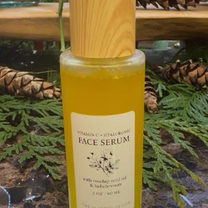 May include: A clear glass bottle of face serum with a wooden cap. The serum is yellow and has a white label with the text "Vitamin C + Hyaluronic Face Serum". The label also lists ingredients and the volume of the product, 2 oz / 60 ml.