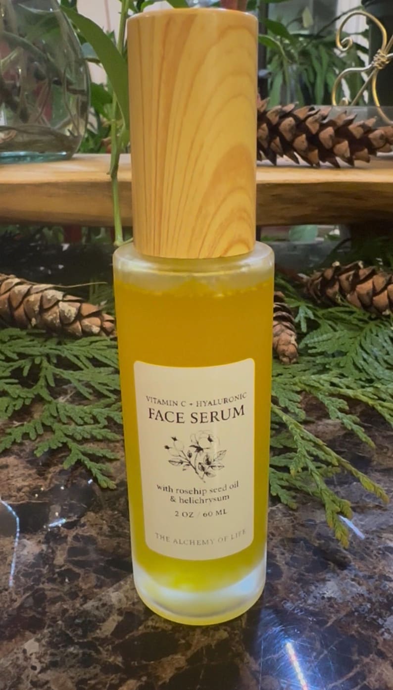 May include: A clear glass bottle of face serum with a wooden cap. The serum is yellow and has a white label with the text "Vitamin C + Hyaluronic Face Serum". The label also lists ingredients and the volume of the product, 2 oz / 60 ml.