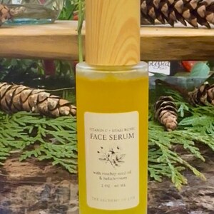 May include: A clear glass face serum bottle with a wooden cap. The bottle contains a yellow liquid and has a label with the text "Vitamin C + Hyaluronic Face Serum". The label also mentions rosehip seed oil and helichrysum.