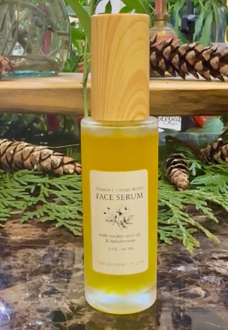 May include: A clear glass face serum bottle with a wooden cap. The bottle contains a yellow liquid and has a label with the text "Vitamin C + Hyaluronic Face Serum". The label also mentions rosehip seed oil and helichrysum.