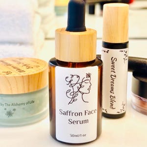 May include: A brown glass bottle of Saffron Face Serum with a wooden cap and black dropper. The label features a line drawing of a woman's face and the text "Saffron Face Serum 30ml/1 oz". A jar of body butter and a roller bottle are also visible.