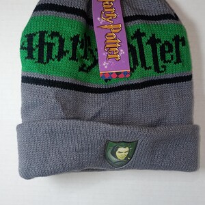 May include: A grey knit beanie featuring a horizontal green stripe with the black text Harry Potter. A purple product tag with the text Harry Potter is attached to the top. The cuff displays a small shield patch featuring a character illustration.