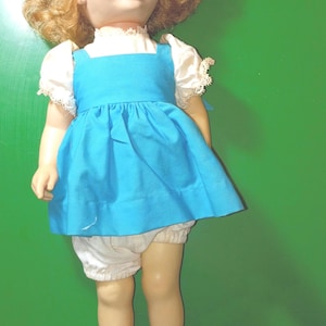 May include: A vintage doll with blonde, curly hair, wearing a blue dress, white bloomers, and white shoes. The doll has a serene expression and is posed against a green background. A classic toy.