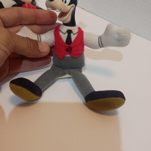 Op de afbeelding: Een Goofy pluche pop met een zwart lichaam, witte handschoenen en een rood vest. De pop heeft een groene hoed en een grijze broek. De schoenen van de pop zijn zwart met een gele zool. Een vintage verzamelobject.