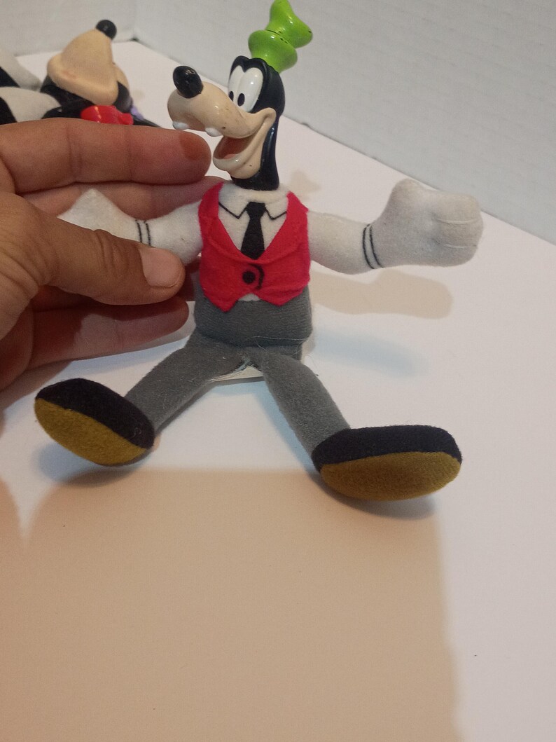 Op de afbeelding: Een Goofy pluche pop met een zwart lichaam, witte handschoenen en een rood vest. De pop heeft een groene hoed en een grijze broek. De schoenen van de pop zijn zwart met een gele zool. Een vintage verzamelobject.