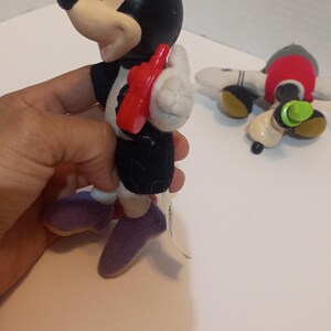 Op de afbeelding: Een Minnie Mouse-beeldje met een rode telefoon, zwarte jurk en paarse schoenen. Het beeldje heeft een zwart hoofd met een paarse strik. Een Mickey Mouse-knuffel staat op de achtergrond.