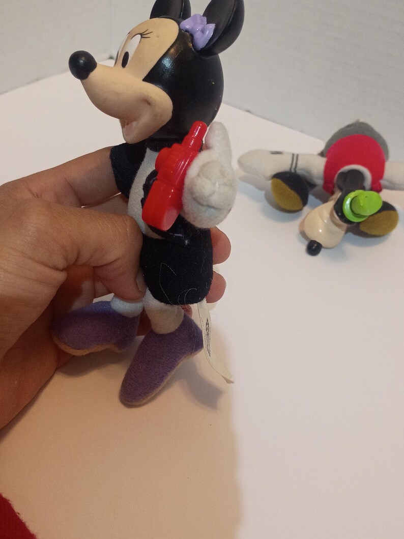 Op de afbeelding: Een Minnie Mouse-beeldje met een rode telefoon, zwarte jurk en paarse schoenen. Het beeldje heeft een zwart hoofd met een paarse strik. Een Mickey Mouse-knuffel staat op de achtergrond.