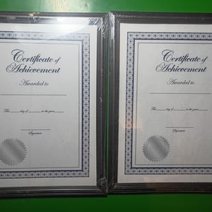 May include: Two framed certificates of achievement. Each certificate has a decorative blue border and the words "Certificate of Achievement" at the top. The frames are black.