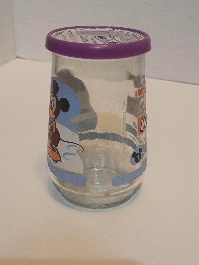 Puede incluir: Un tarro de cristal transparente con tapa morada y un dibujo de Mickey Mouse. El tarro tiene forma cil&iacute;ndrica y un borde azul claro con el texto "7-Up Mickey Mouse". El tarro est&aacute; vac&iacute;o.