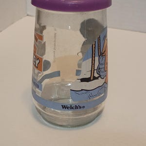 Puede incluir: Un tarro de cristal transparente con tapa morada. El tarro tiene una banda azul en la parte inferior con el logotipo de "Welch's". El tarro tiene un gr&aacute;fico de dibujos animados en el lateral. El tarro es cil&iacute;ndrico y parece vac&iacute;o.