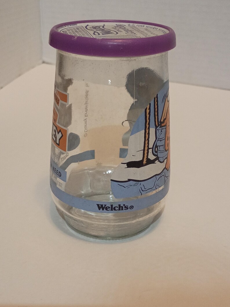 Puede incluir: Un tarro de cristal transparente con tapa morada. El tarro tiene una banda azul en la parte inferior con el logotipo de "Welch's". El tarro tiene un gr&aacute;fico de dibujos animados en el lateral. El tarro es cil&iacute;ndrico y parece vac&iacute;o.