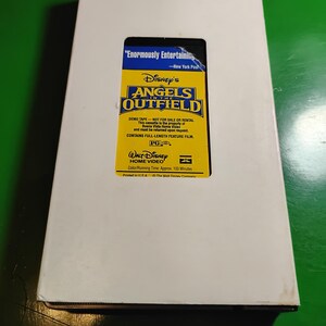 May include: A white cardboard box with a rectangular cutout revealing a yellow VHS tape. The tape is labeled "Disney's Angels in the Outfield" and includes text from the New York Post, "Enormously Entertaining."