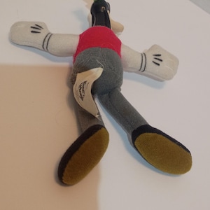 Op de afbeelding: Een pluche speelgoed van Goofy, een Disney-stripfiguur. Het speelgoed heeft een rood vest, een grijze broek, witte handschoenen met zwarte details en een groene hoed. De voeten zijn geel met zwarte zolen.