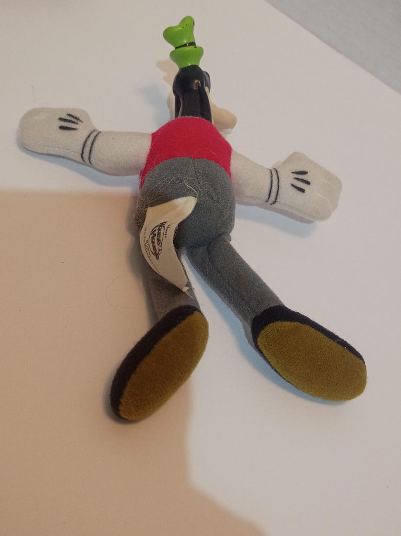 Op de afbeelding: Een pluche speelgoed van Goofy, een Disney-stripfiguur. Het speelgoed heeft een rood vest, een grijze broek, witte handschoenen met zwarte details en een groene hoed. De voeten zijn geel met zwarte zolen.