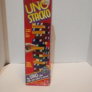 May include: A red box for the game UNO Stacko. The box features the game's logo in yellow and the game's name. The box includes the text "For ages 8 and older. For 2 or more players." The game includes 51 blocks, an UNO cube, and a loading tray.