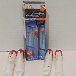 May include: A set of four white battery-operated LED hanging candles with red wax-like drips at the top. The packaging box displays the text Celebrate! Halloween LED Flickering Flame White Hanging Candles, 7 IN (17.7 cm) CANDLE HEIGHT.
