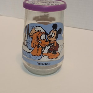 Puede incluir: Un tarro de vidrio transparente con tapa morada que presenta un gr&aacute;fico de dibujos animados de Mickey Mouse y Pluto. El tarro tiene una banda azul con el logotipo de "Welch's". Los personajes est&aacute;n en una escena invernal.