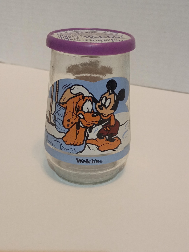 Puede incluir: Un tarro de vidrio transparente con tapa morada que presenta un gr&aacute;fico de dibujos animados de Mickey Mouse y Pluto. El tarro tiene una banda azul con el logotipo de "Welch's". Los personajes est&aacute;n en una escena invernal.