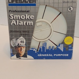 May include: A Lifesaver Professional Smoke Alarm in its original box. The box is gray and white with the words "Smoke Alarm" in large, bold letters. The alarm has a white, circular design with red accents and the text "General Purpose".